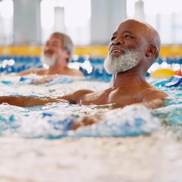 Two older men with beards enjoying swimming in a pool, surrounded by blue water and lane dividers in a well-lit indoor environment.