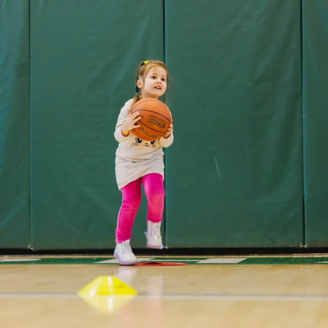 A young girl in pink leggings jumps while holding a basketball, with a boy nearby also ready for youth basketball practice in a gym with green padded walls.