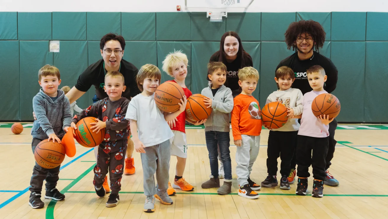 A group of young children holding basketballs poses with three adult coaches on an indoor youth basketball court.