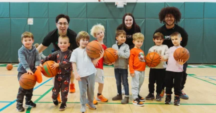 A group of young children holding basketballs poses with three adult coaches on an indoor youth basketball court.