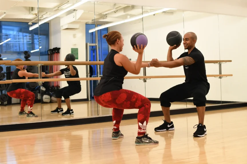 Two people follow a 2026 fitness routine in a gym studio, squatting with medicine balls and facing each other in front of mirrors.