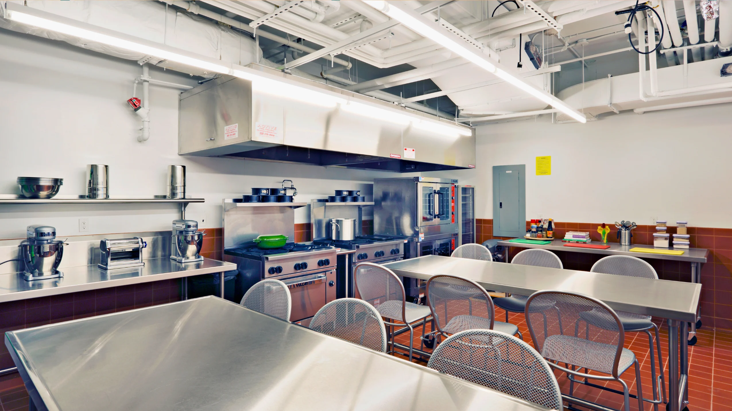 Commercial kitchen with stainless steel tables, metal chairs, industrial stoves, cookware, and utensils under bright overhead lighting.