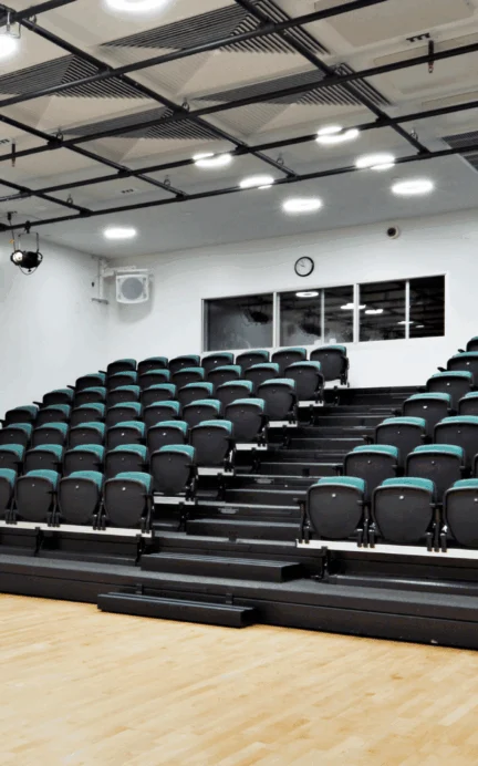 A small auditorium with black and teal tiered seating, wooden floor, white walls, ceiling lights, and a clock above a window at the back.
