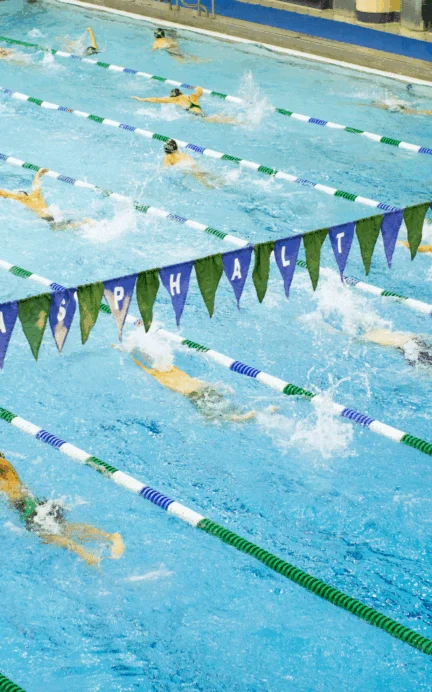 Swimmers compete in multiple lanes of an indoor swimming pool, separated by lane dividers, with water splashing as they race.