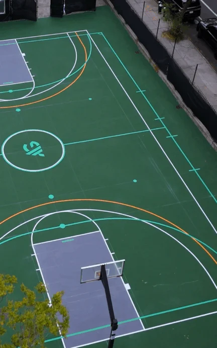 Aerial view of an empty outdoor basketball court with clear markings, surrounded by a fence, trees, and a nearby street with parked cars.